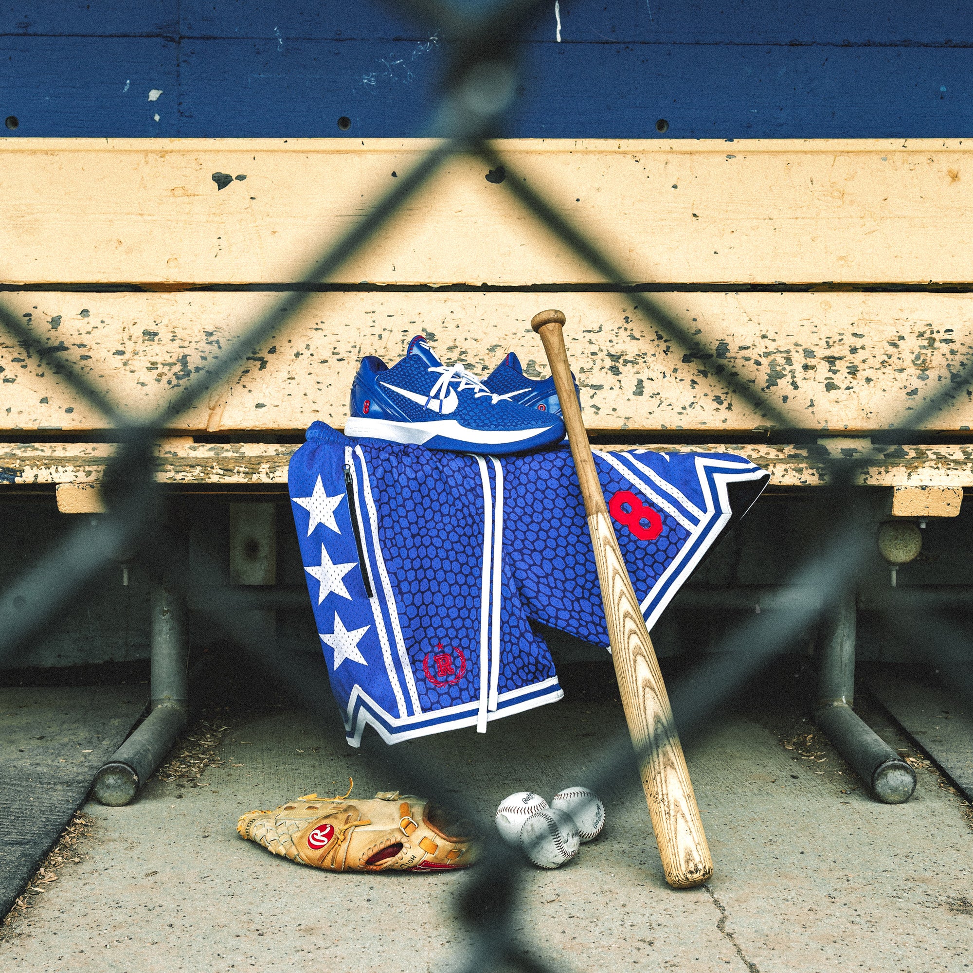 Heavyweight mesh basketball shorts with mamba snakeskin pattern, white stars, red embroidered number 8, rib knit trim, and long drawstrings—hanging on dugout bench with baseball glove, bat, and Nike Kobe 6 Protro Game Royal sneakers
