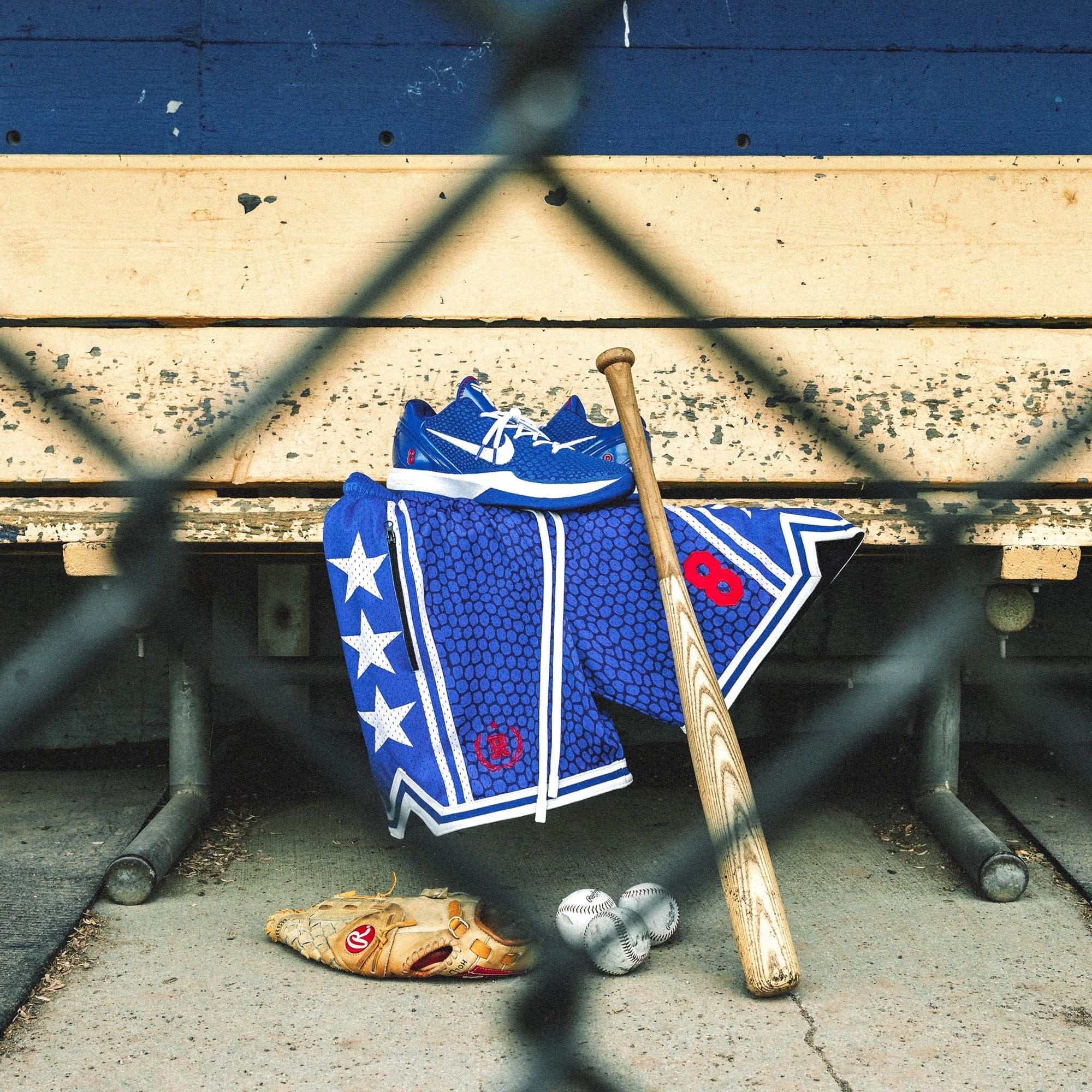 Heavyweight mesh basketball shorts with mamba snakeskin pattern, white stars, red embroidered number 8, rib knit trim, and long drawstrings—hanging on dugout bench with baseball glove, bat, and Nike Kobe 6 Protro Game Royal sneakers