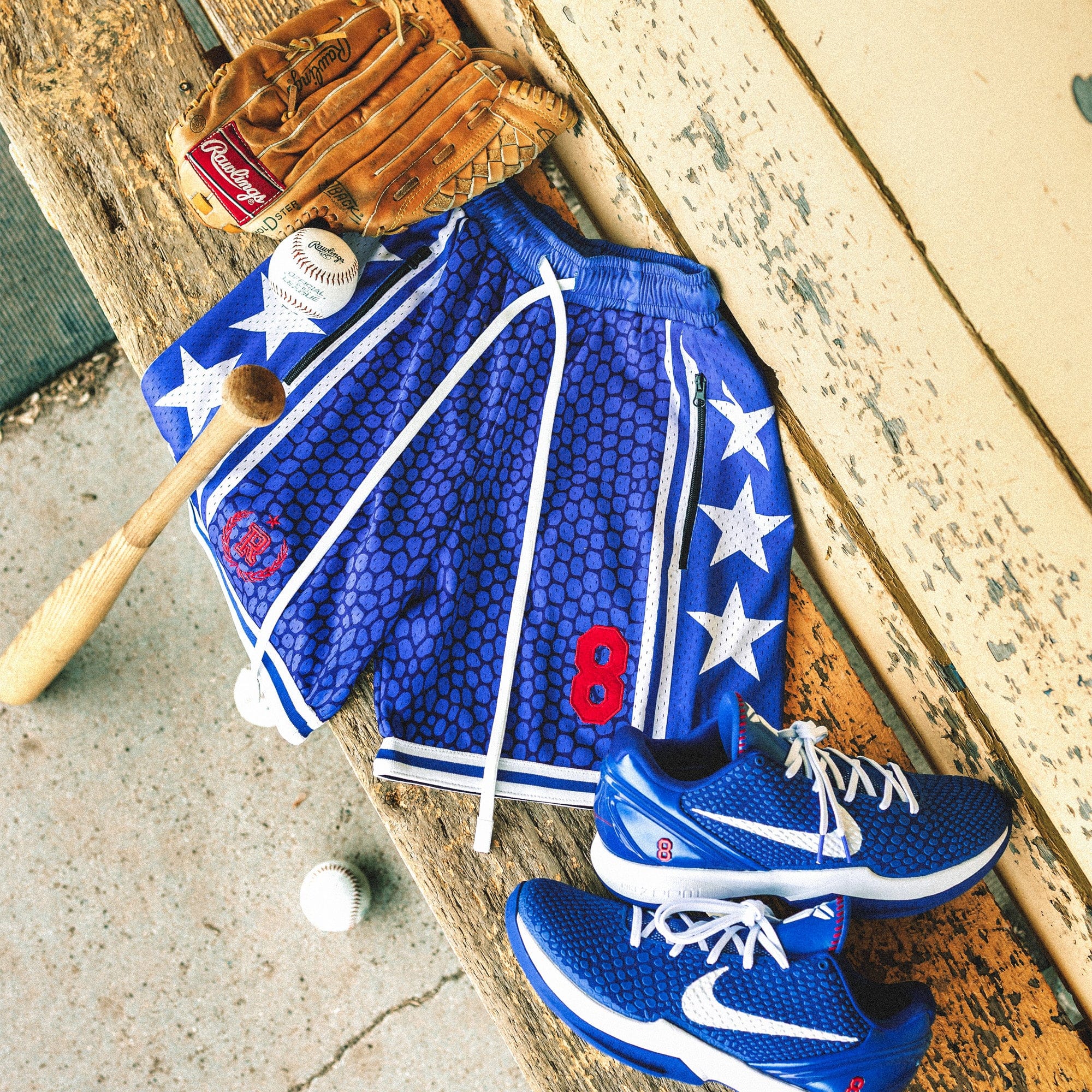 Heavyweight mesh basketball shorts with mamba snakeskin pattern, white stars, red embroidered number 8, rib knit trim, and long drawstrings—hanging on dugout bench with baseball glove, bat, and Nike Kobe 6 Protro Game Royal sneakers