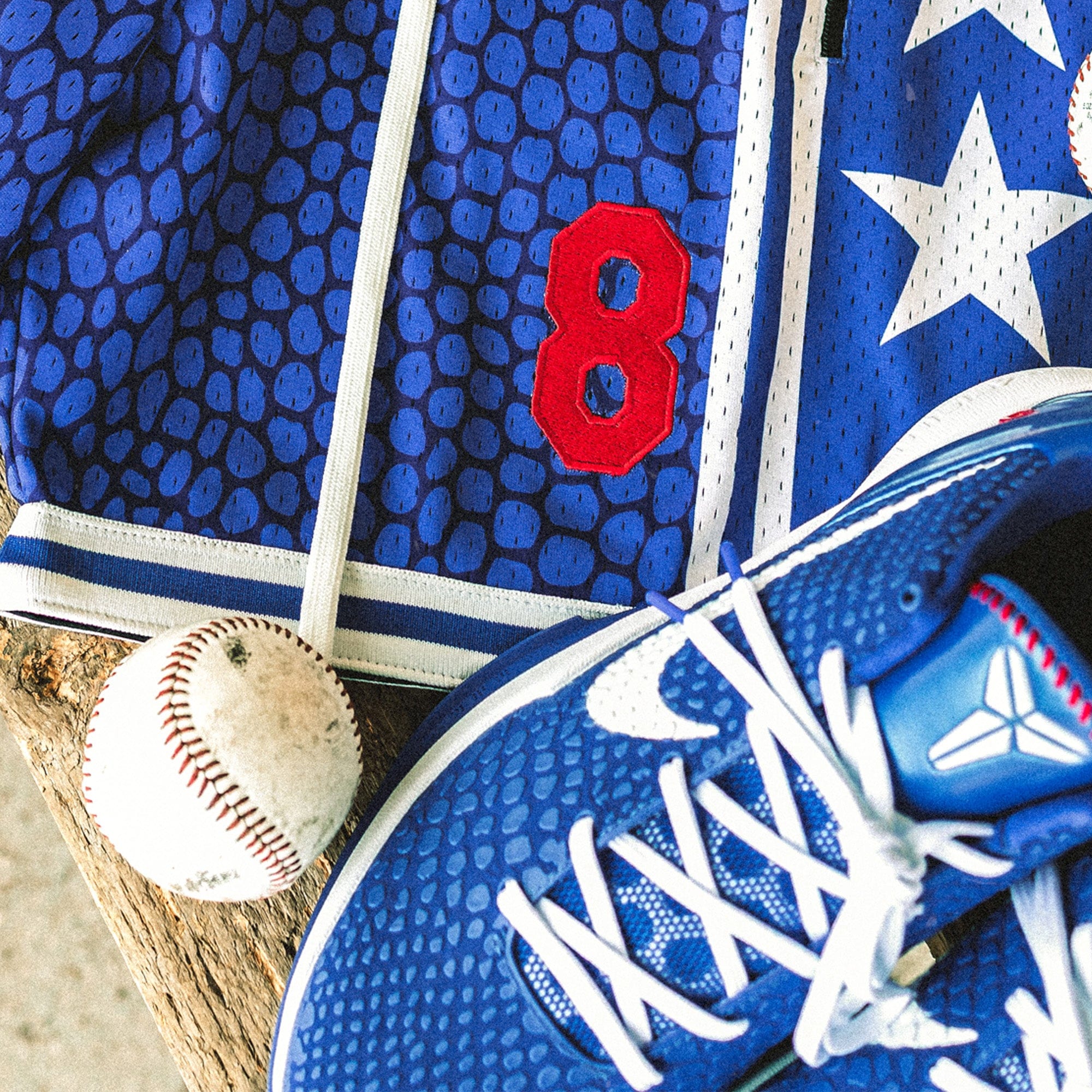 Heavyweight mesh basketball shorts with mamba snakeskin pattern, white stars, red embroidered number 8, rib knit trim, and long drawstrings—hanging on dugout bench with baseball glove, bat, and Nike Kobe 6 Protro Game Royal sneakers