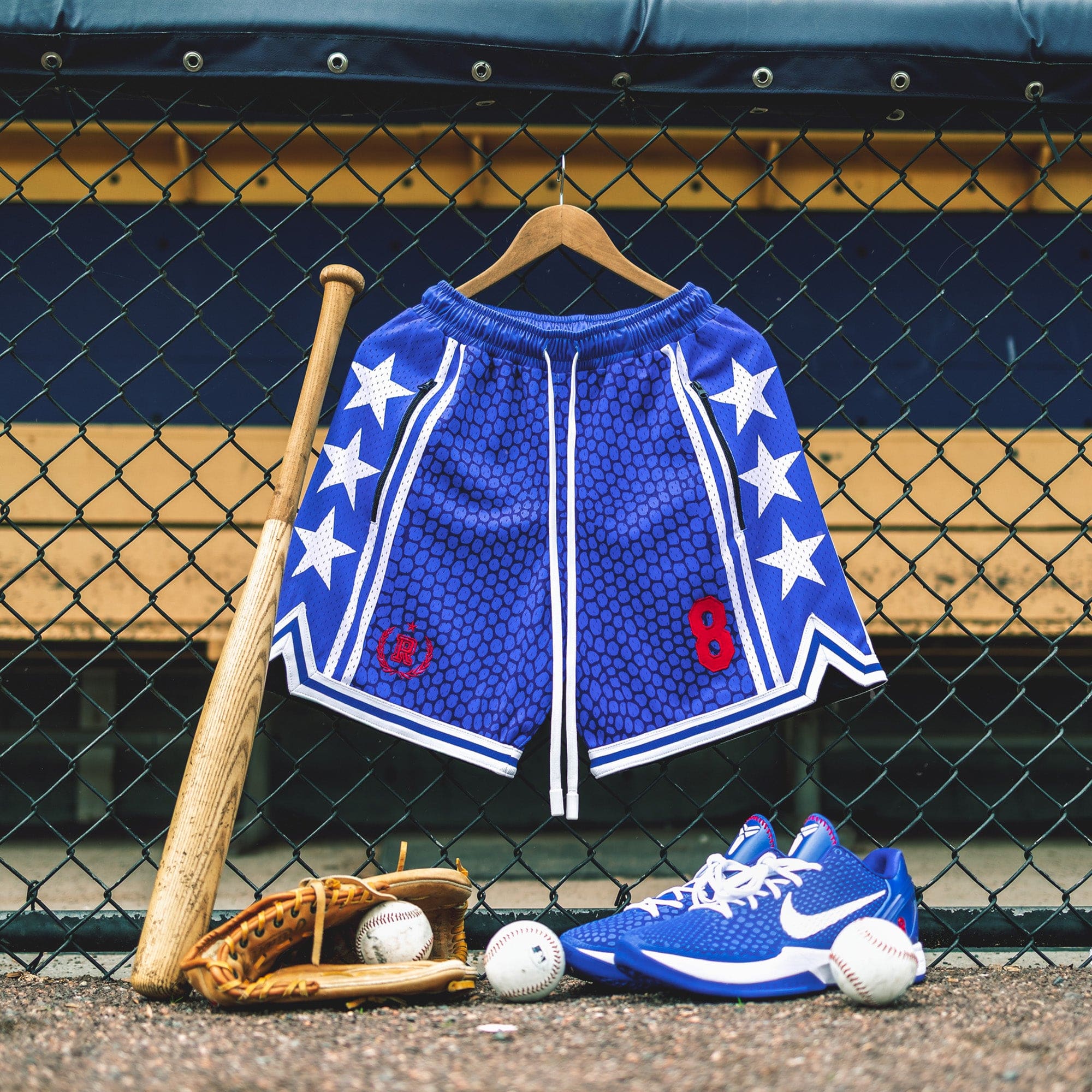 Heavyweight mesh basketball shorts with mamba snakeskin pattern, white stars, red embroidered number 8, rib knit trim, and long drawstrings—hanging on dugout fence with baseball glove, bat, and Nike Kobe 6 Protro Game Royal basketball sneakers