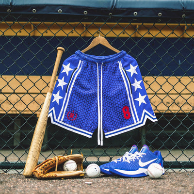Heavyweight mesh basketball shorts with mamba snakeskin pattern, white stars, red embroidered number 8, rib knit trim, and long drawstrings—hanging on dugout fence with baseball glove, bat, and Nike Kobe 6 Protro Game Royal basketball sneakers